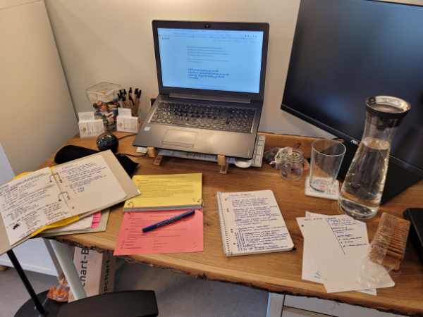 Ein hölzerner Schreibtisch, auf dem verschiedene Unterlagen und handschriftliche Notizen vor einem Laptop ausgebreitet liegen.